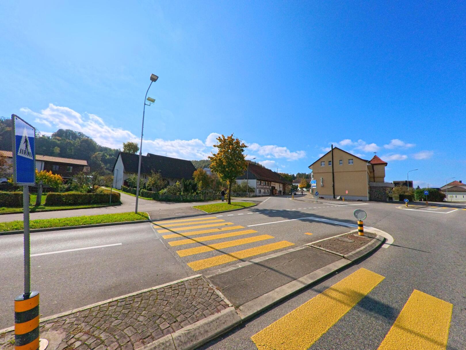 Ein Fussgaengerstreifen fuehrt in Leibstadt (5325, Kanton Aargau) ueber eine breite Strasse, gesaeumt von Wohnhaeusern und einem markanten Gebaeude im Hintergrund.