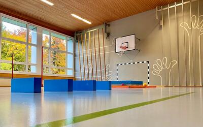 In einer Sporthalle in Nesslau (9650, Kanton St. Gallen) stehen blaue Turnelemente vor Sprossenwänden, Basketballkorb und Hallentor.