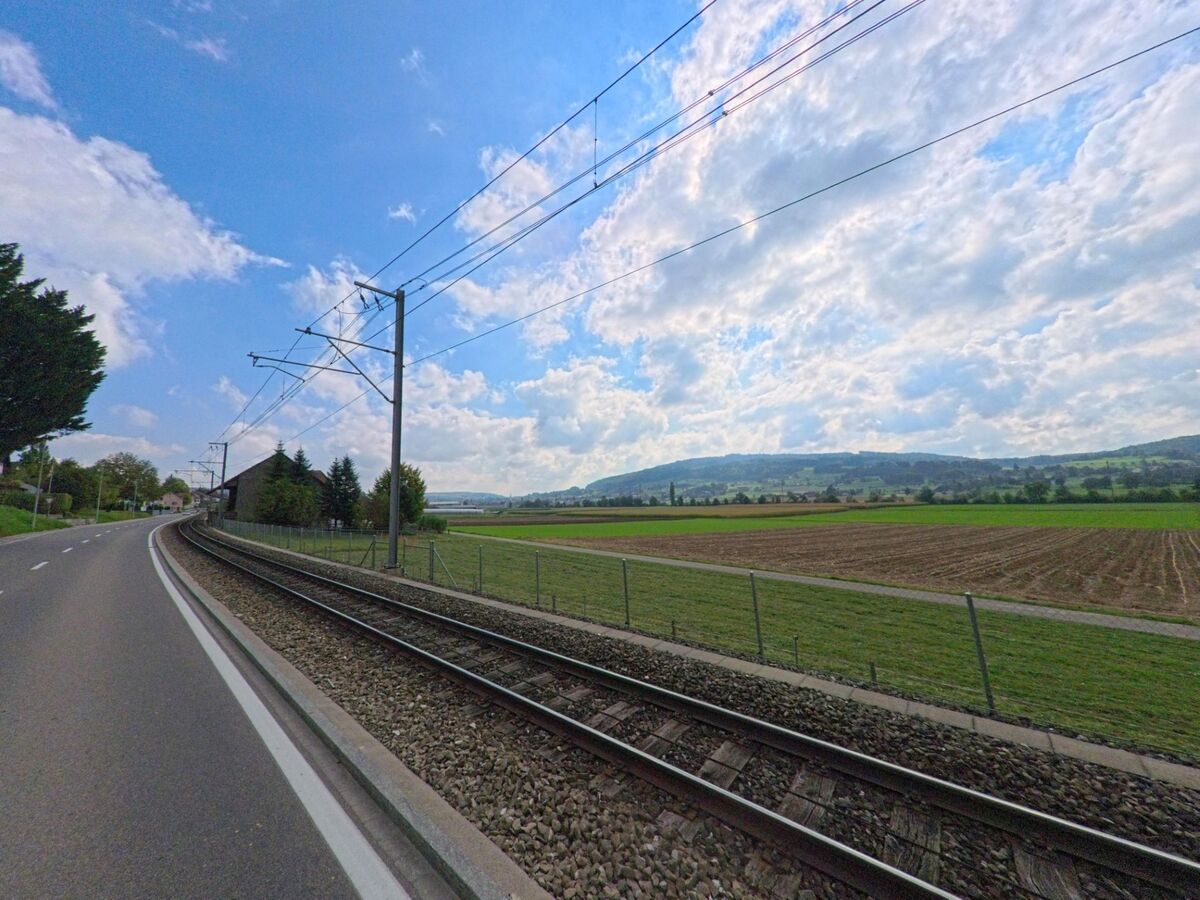 Die Bahnlinie verläuft in Leimbach (5733, Kanton Aargau) parallel zur Hauptstrasse und führt durch eine weite Landschaft mit Feldern und sanften Hügeln.
