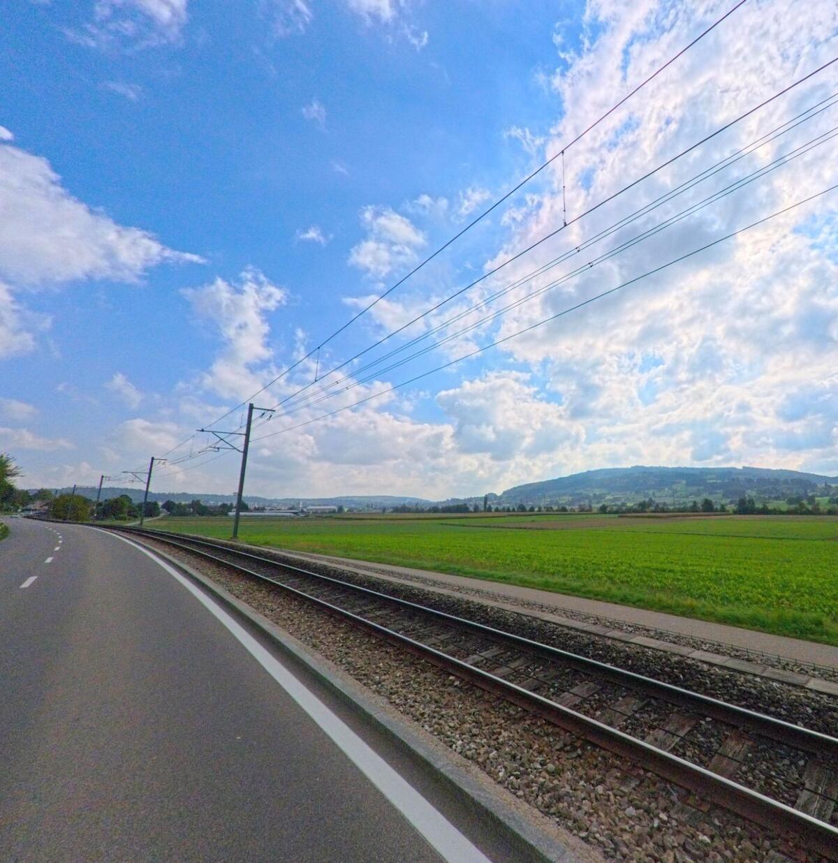 Zwischen der Bahnlinie und der Hauptstrasse in Leimbach (5733, Kanton Aargau) erstrecken sich grüne Felder mit Blick auf die Hügellandschaft im Hintergrund.