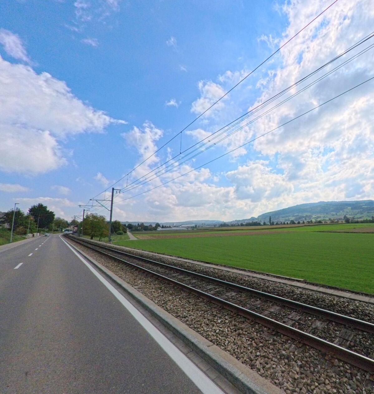 In Leimbach (5733, Kanton Aargau) verläuft die Bahnstrecke parallel zur Strasse, neben der sich weite grüne Felder bis zum Hügelgebiet erstrecken.