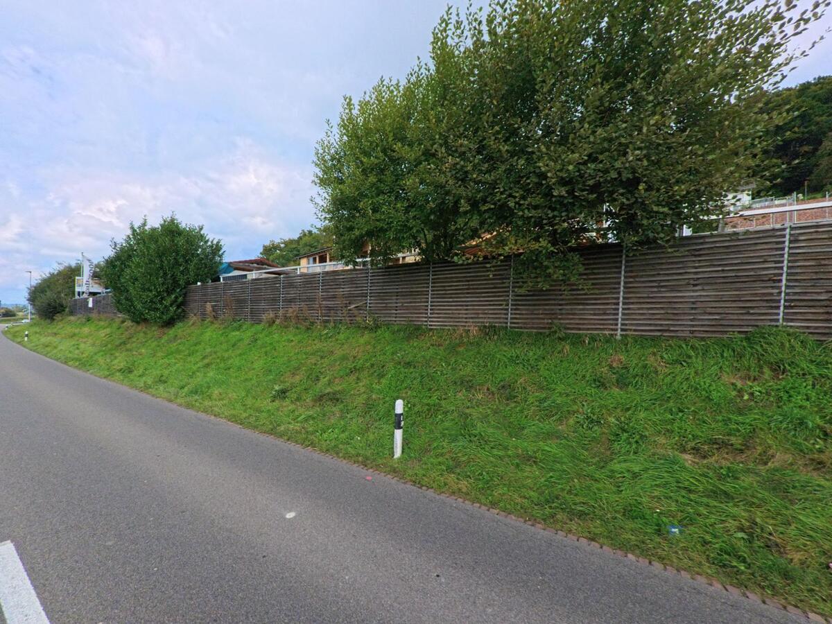 Entlang der ansteigenden Strasse in Leimbach (5733, Kanton Aargau) verläuft ein hoher Holzzaun, hinter dem Wohnhäuser und Bäume zu sehen sind.