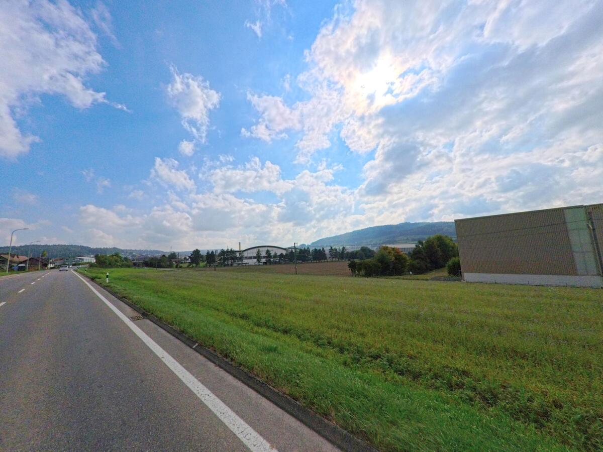 Am Dorfrand von Leimbach (5733, Kanton Aargau) führt eine breite Strasse an Feldern und Gewerbebauten vorbei, im Hintergrund sind Hügel und Wald zu sehen.