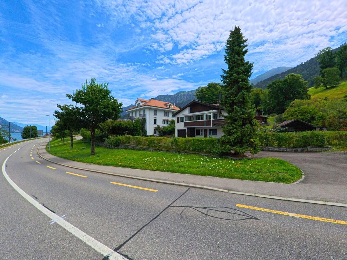 An einer sanft geschwungenen Strasse in Leissigen (3706, Kanton Bern) stehen ein weisses Herrenhaus und ein modernes Wohnhaus vor bewaldeten Berghaengen.