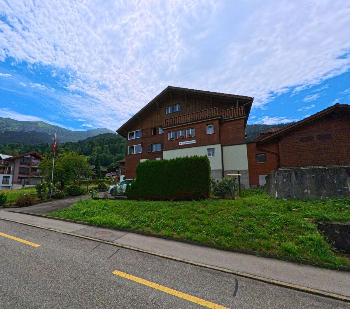 Ein grosses Wohnhaus mit Holzfassade steht in Leissigen (3706, Kanton Bern) am Rand einer gepflegten Gruenflaeche vor bewaldeten Bergen.