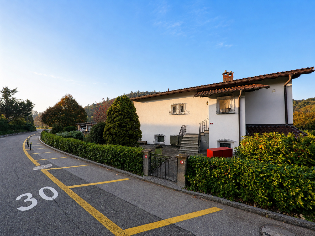 An einer verkehrsberuhigten Strasse in Lema (6981, Kanton Tessin) steht ein helles Wohnhaus hinter gepflegten Hecken und einem kleinen Vorgarten.