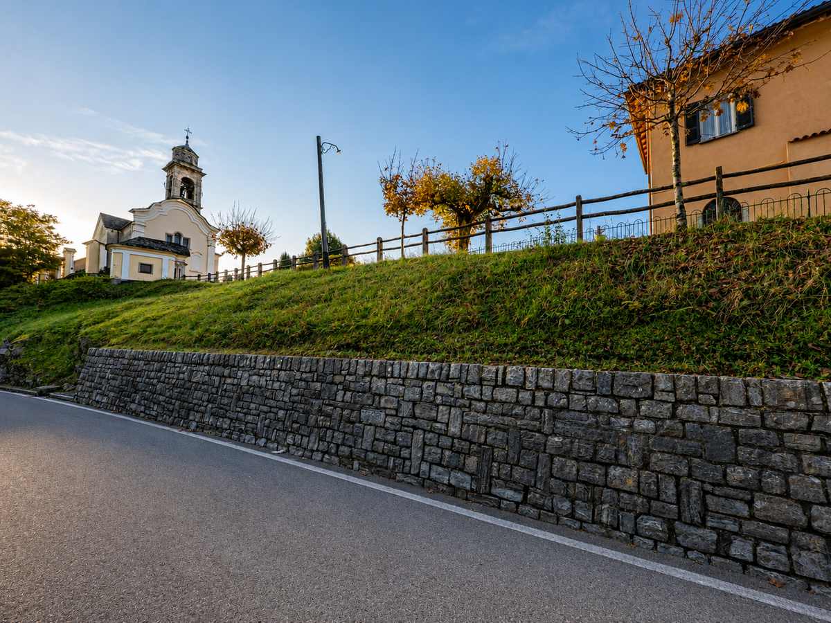 Oberhalb einer von einer Natursteinmauer gesäumten Strasse steht in Lema (6992, Kanton Tessin) eine kleine Kirche mit Glockenturm auf einem begrünten Hang.