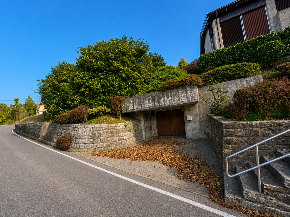 An einer ansteigenden Strasse in Lema (6997, Kanton Tessin) liegt eine in den Hang integrierte Garage zwischen Natursteinmauern und dicht bepflanzten Gartenflächen.