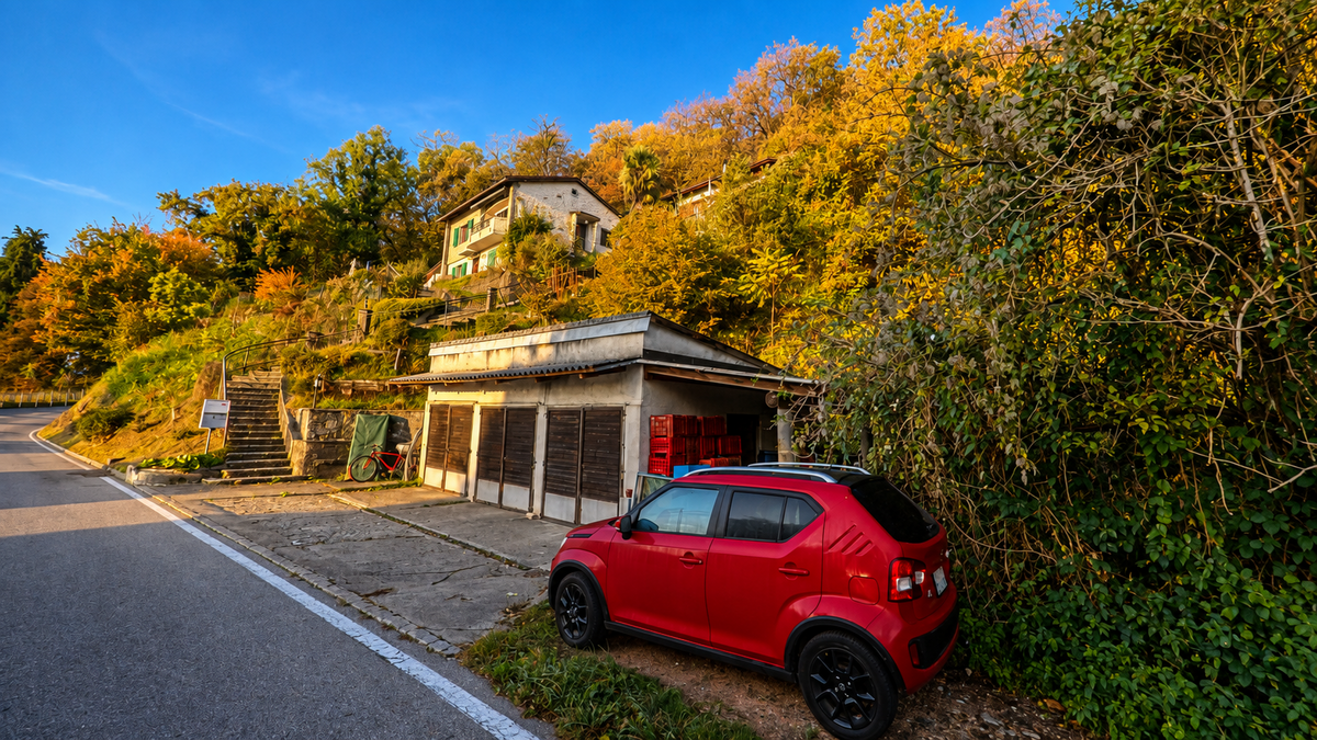 An einer kurvigen Strasse in Lema (6997, Kanton Tessin) liegen eine Garage und ein Wohnhaus am bewaldeten Hang zwischen herbstlich gefärbten Bäumen.