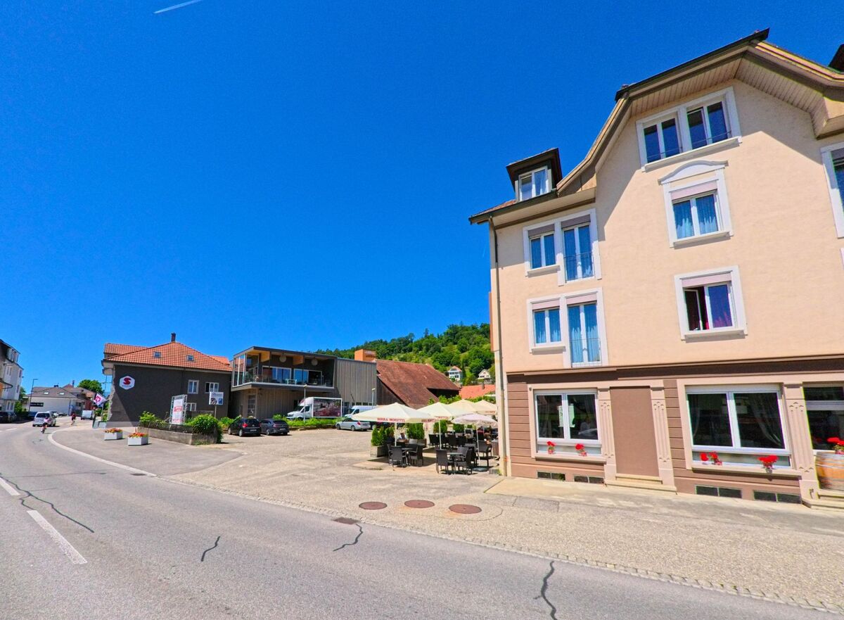 In Lengnau (2543, Kanton Bern) führt eine belebte Straße entlang eines Hauses mit einem Café und sonnigen Tischen im Vordergrund.