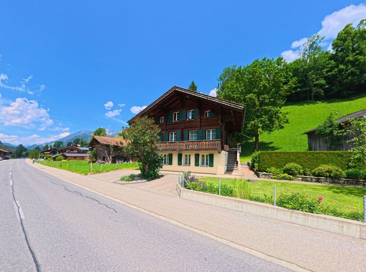 In Lenk (3775, Kanton Bern) steht ein traditionelles Wohnhaus mit gruenen Fensterlaeden direkt an einer ruhig verlaufenden Strasse vor einem sonnigen Hang.