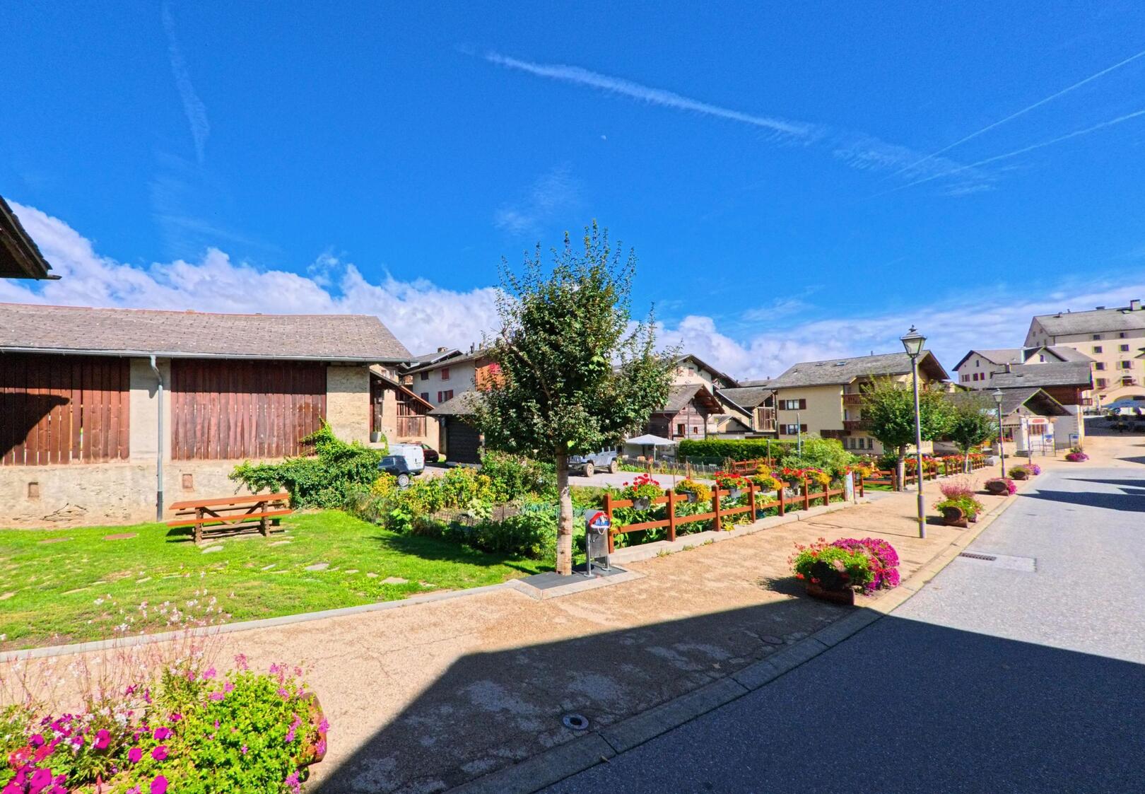 In Lens (3978, Kanton Wallis) zeigt sich eine gepflegte Strasse mit Blumenbeeten, Baenken und traditionellen Haeusern im Hintergrund.
