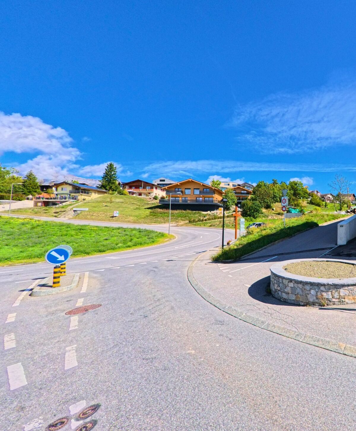 Eine kurvige Strasse fuehrt in Lens (3978, Kanton Wallis) an einer Verkehrsinsel vorbei hinauf zu Haeusern am Hang.
