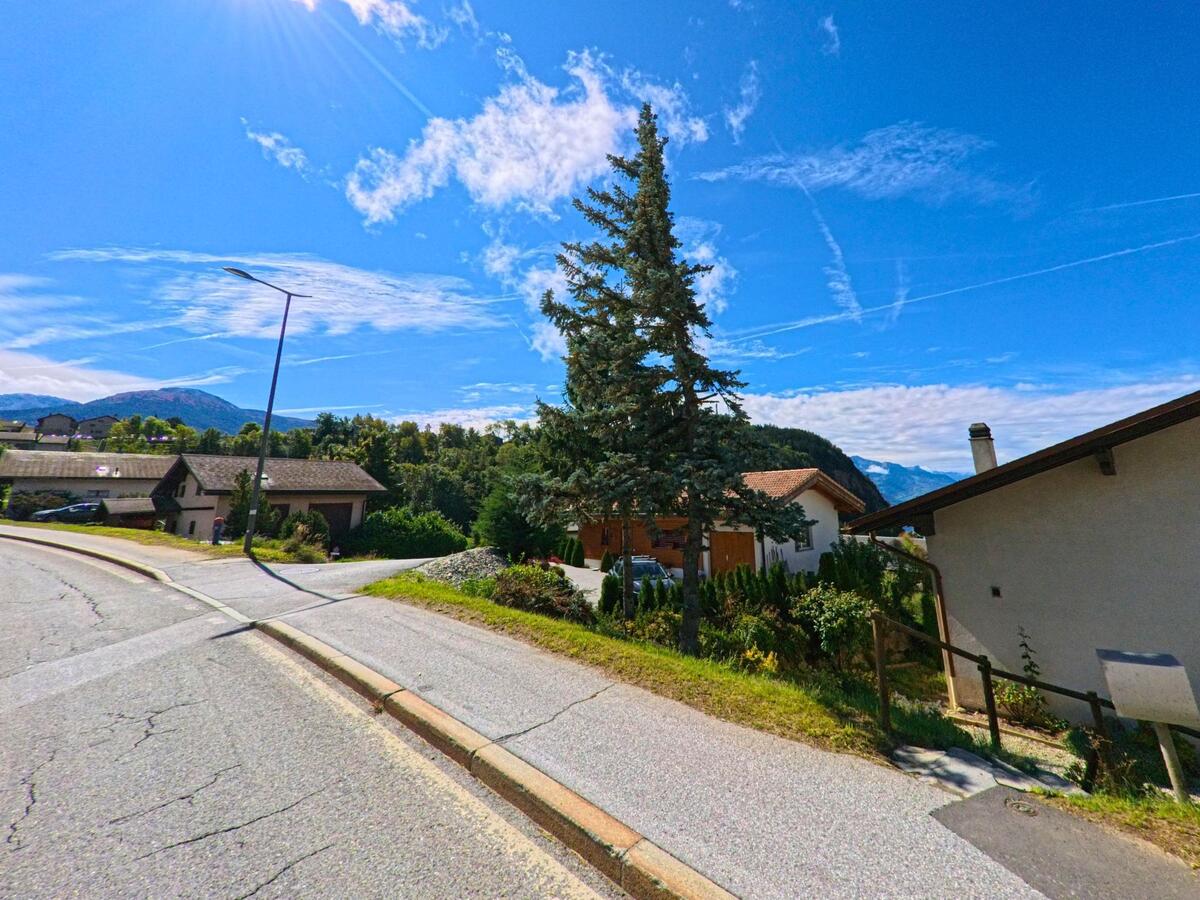 In Lens (3978, Kanton Wallis) verlaeuft eine Nebenstrasse an Einfamilienhaeusern vorbei, flankiert von einem grossen Nadelbaum.