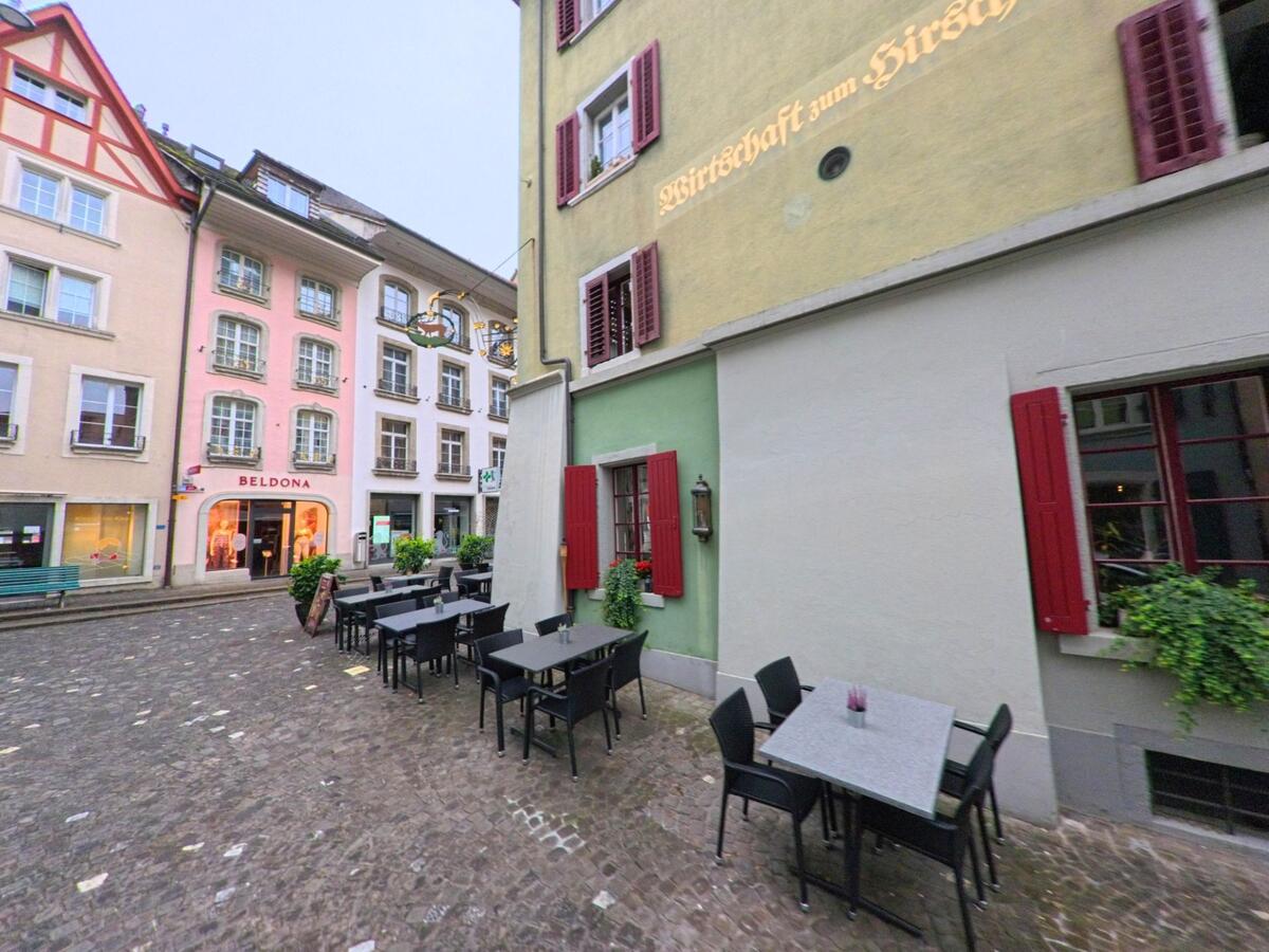 In der Altstadt von Lenzburg (5600, Kanton Aargau) stehen entlang einer gepflasterten Gasse farbige Häuser und ein Restaurant mit Aussensitzplätzen.