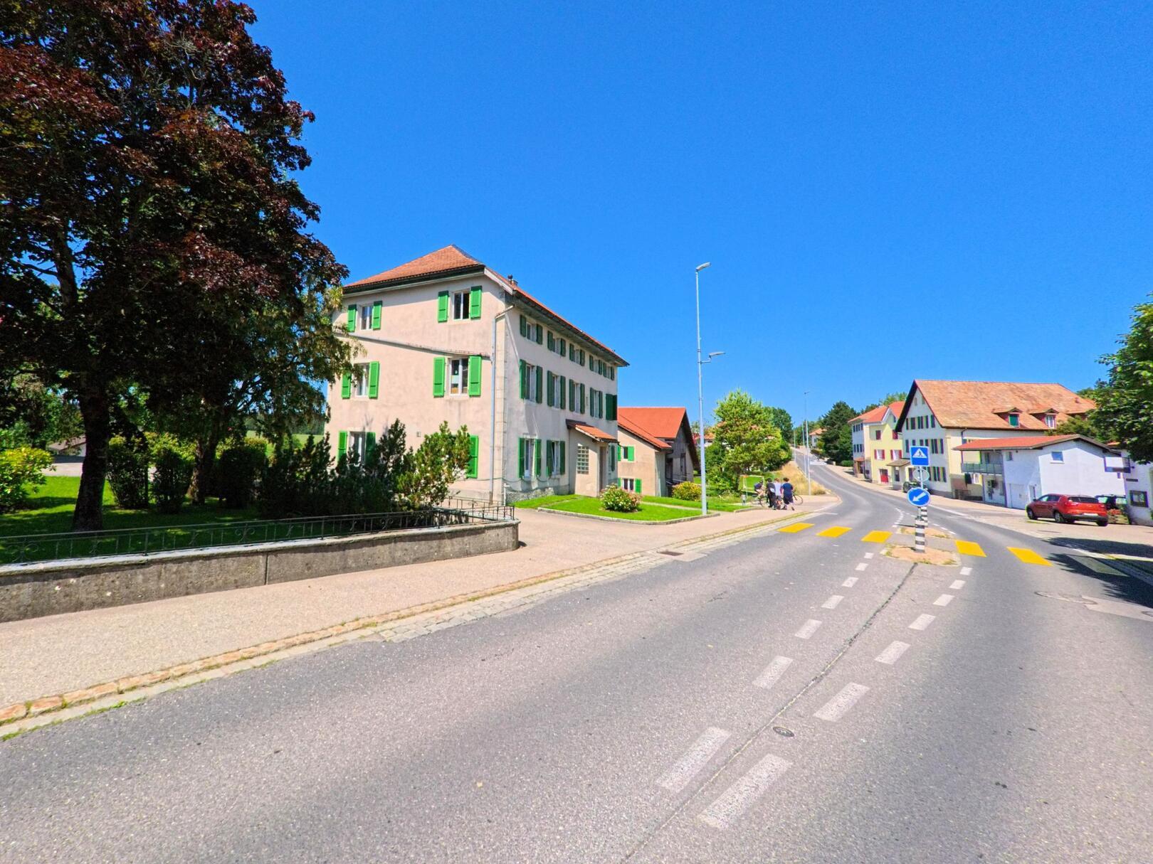 Entlang der Hauptstrasse in Les Bois (2336, Kanton Jura) stehen gepflegte Haeuser mit grünen Fensterlaeden und vorgelagerten Gaerten.
