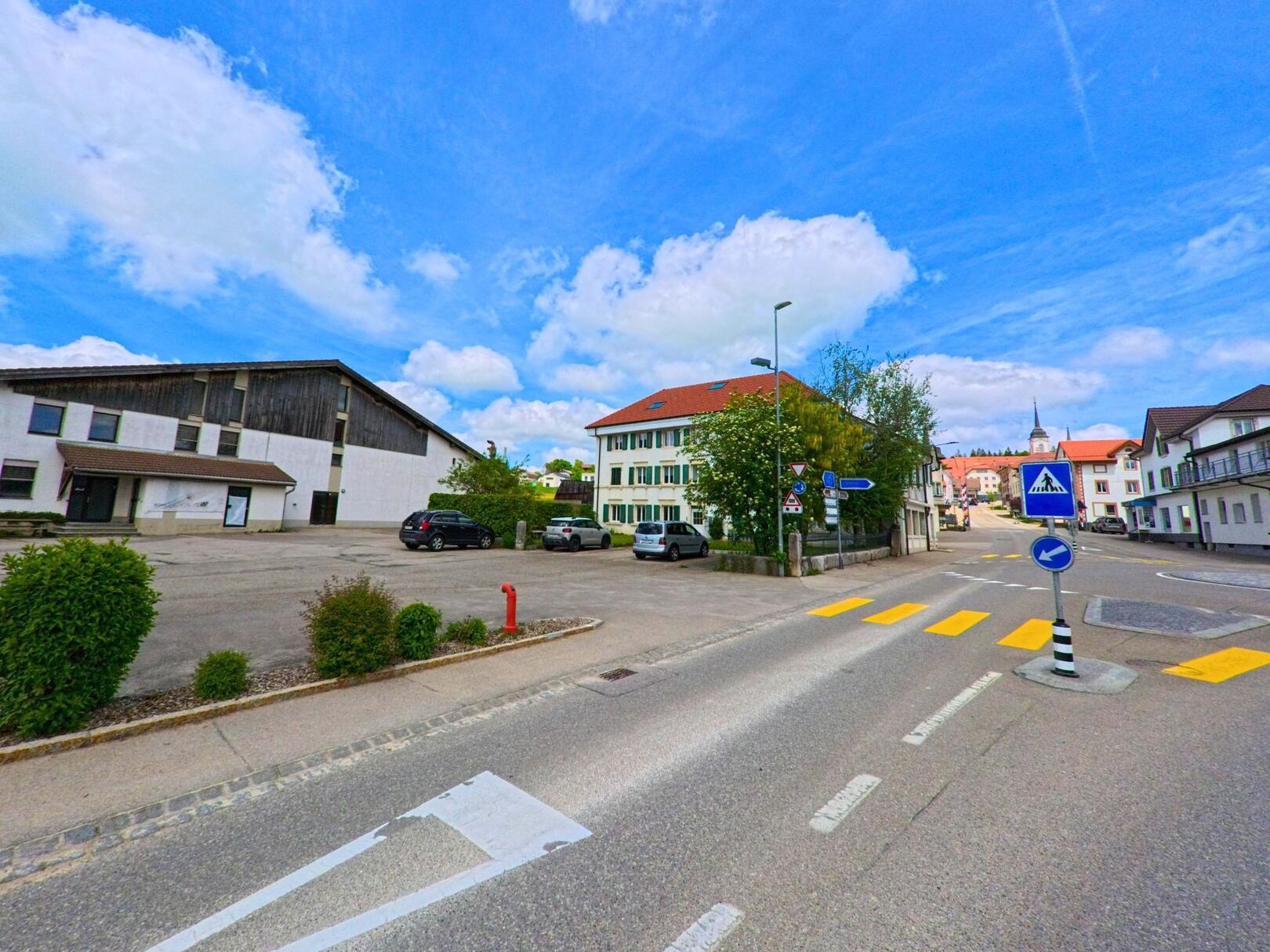 In Les Breuleux (2345, Kanton Jura) zeigt die Strasse eine belebte Kreuzung mit gut gepflegten Gebäuden und einem klaren blauen Himmel.