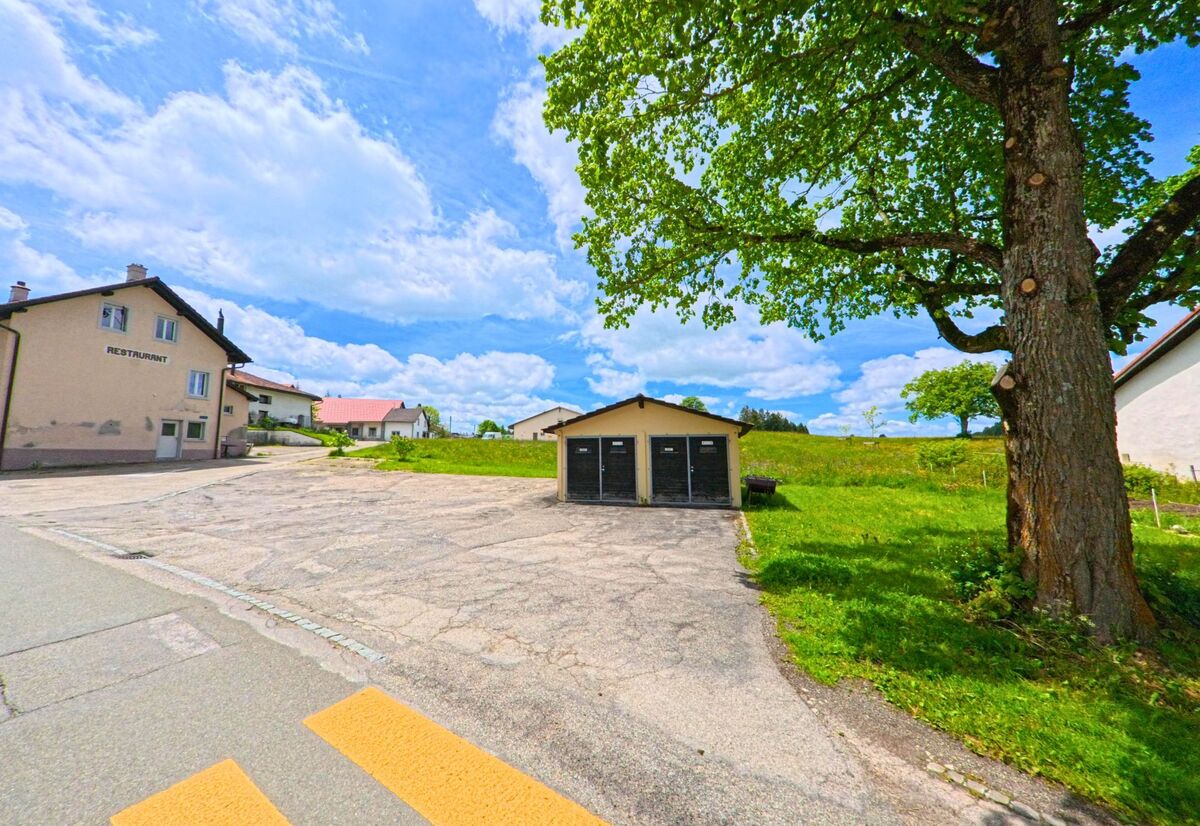 In Les Genevez (2714, Kanton Jura) zeigt sich ein Restaurantgebäude und eine Garage unter einem großen Baum, während die Wiese und der Himmel in leuchtenden Farben erstrahlen.