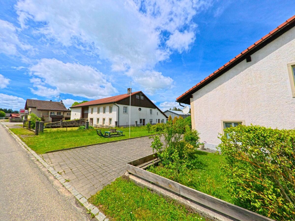 In Les Genevez (2714, Kanton Jura) verläuft eine Straße an Wohngebäuden und einem gepflasterten Gehweg vorbei, während der Himmel in lebendigen Blau- und Weißtönen erstrahlt.