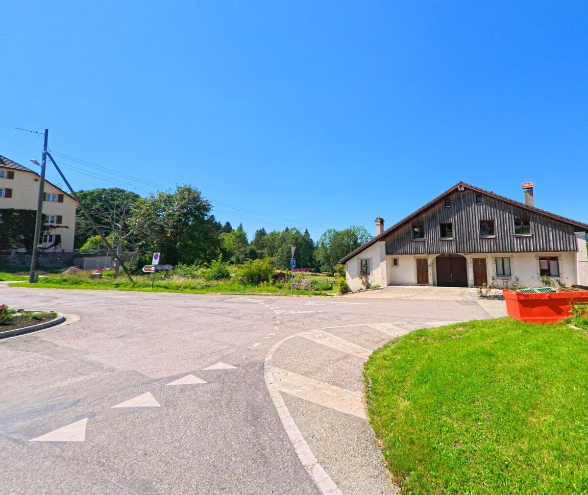 An einer Strassenkreuzung in Les Planchettes (2325, Kanton Neuenburg) steht ein grosses Bauernhaus mit Holzfassade und roten Baucontainern davor.