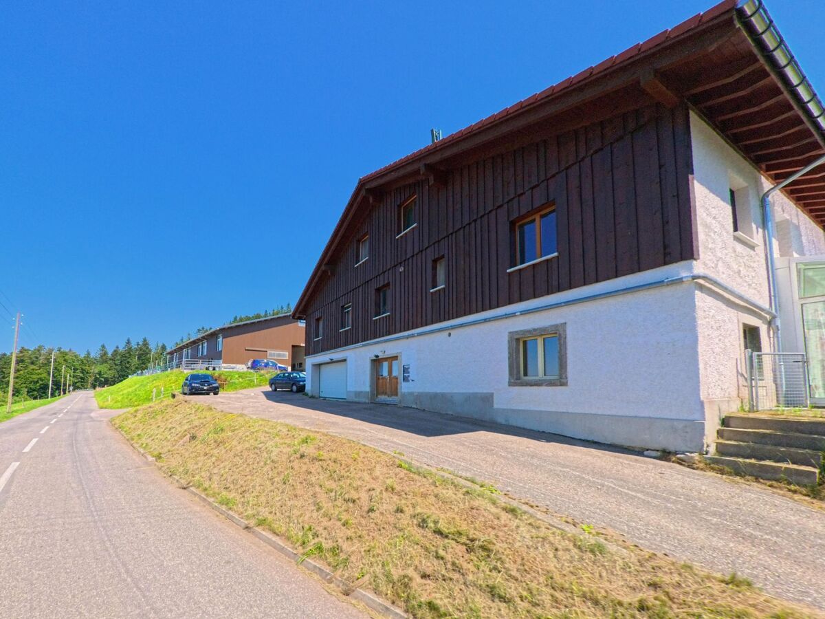 Ein Wohn- und Wirtschaftsgebaeude mit Holzfassade steht in Les Planchettes (2325, Kanton Neuenburg) an einer Landstrasse mit Blick auf Felder und Wald.