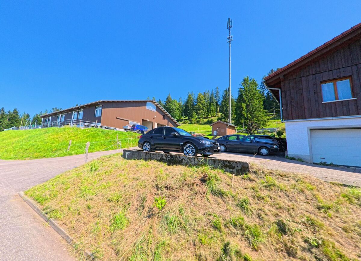Neben einem braunen Hallengebaeude in Les Planchettes (2325, Kanton Neuenburg) stehen mehrere geparkte Autos auf einer leicht ansteigenden Flaeche.