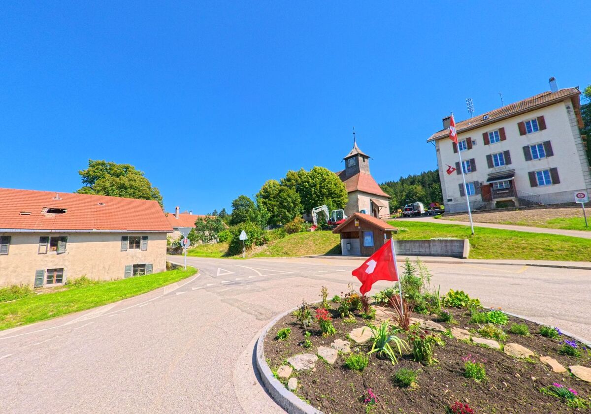 Ein bepflanzter Kreisel mit Schweizer Fahne liegt in Les Planchettes (2325, Kanton Neuenburg) vor dem Gemeindehaus und der Kirche mit Uhrturm.