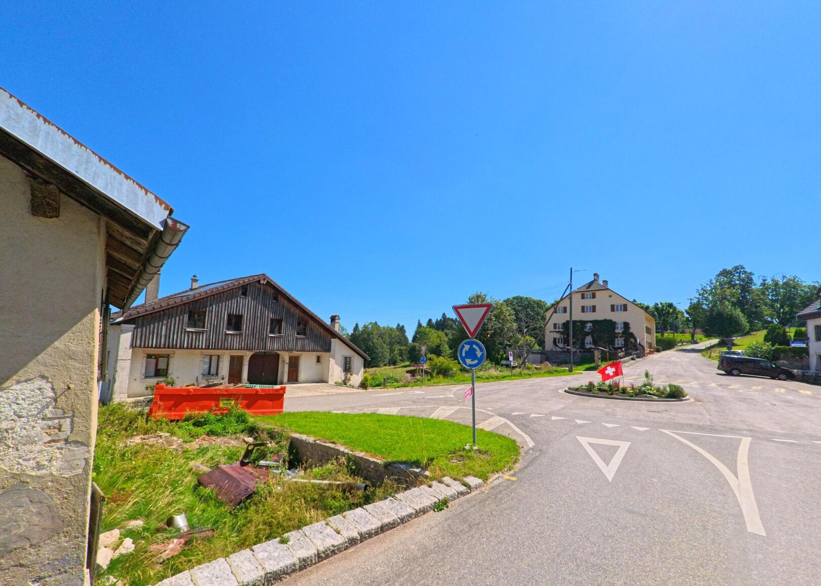 Ein kleiner Kreisverkehr mit Schweizer Fahne liegt in Les Planchettes (2325, Kanton Neuenburg) zwischen Wohnhaeusern und landwirtschaftlichen Gebaeuden.