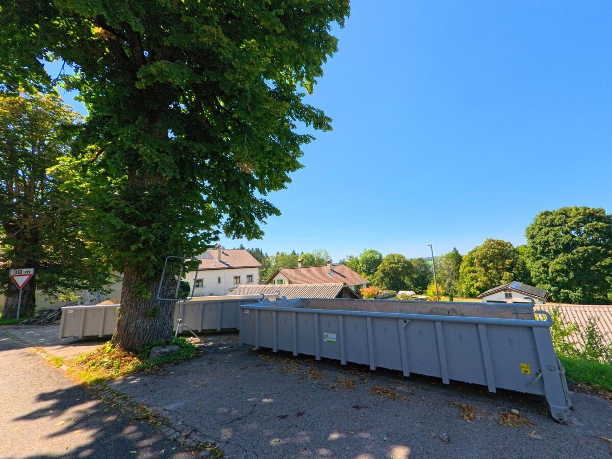 Am Strassenrand in Les Planchettes (2325, Kanton Neuenburg) stehen mehrere grosse Metallcontainer im Schatten eines Baumes.