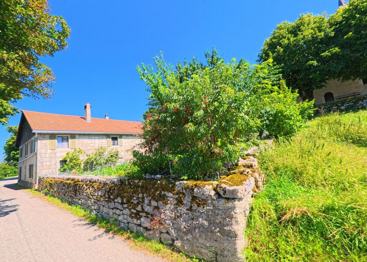 Ein traditionelles Steinhaus mit rotem Ziegeldach steht in Les Planchettes (2325, Kanton Neuenburg) an einer schmalen Strasse, umgeben von Garten und Steinmauer.