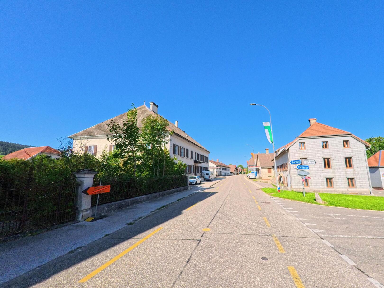 In Les Verrieres (2126, Kanton Neuenburg) fuehrt eine leicht ansteigende Dorfstrasse zwischen traditionellen Wohnhaeusern in Richtung Ortszentrum.