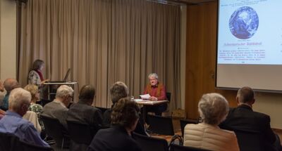 In Horgen (8810, Kanton Zuerich) verfolgt ein Publikum eine Lesung, bei der ein Mann am Tisch vortraegt, waehrend eine Frau am Laptop die Projektion auf der Leinwand steuert.