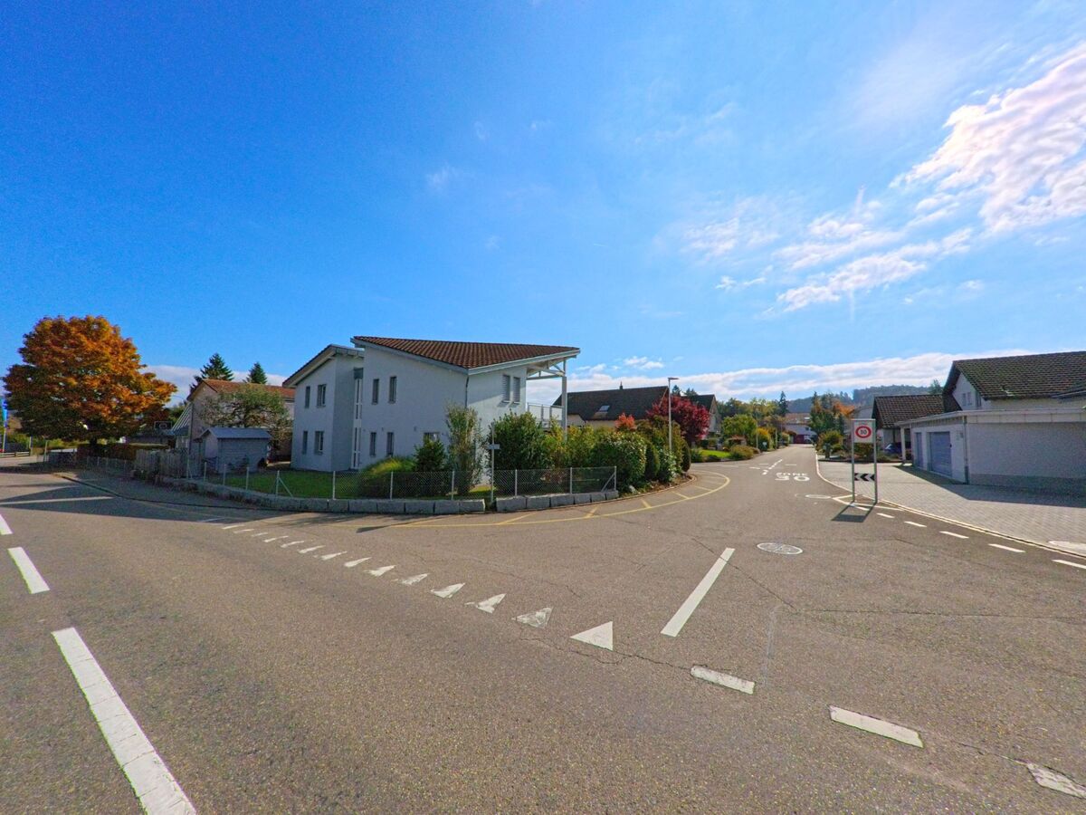 Eine ruhige Quartierstrasse in Leuggern (5316, Kanton Aargau) führt zwischen modernen Einfamilienhäusern mit gepflegten Gärten hindurch, während herbstliche Bäume Farbe ins Bild bringen.
