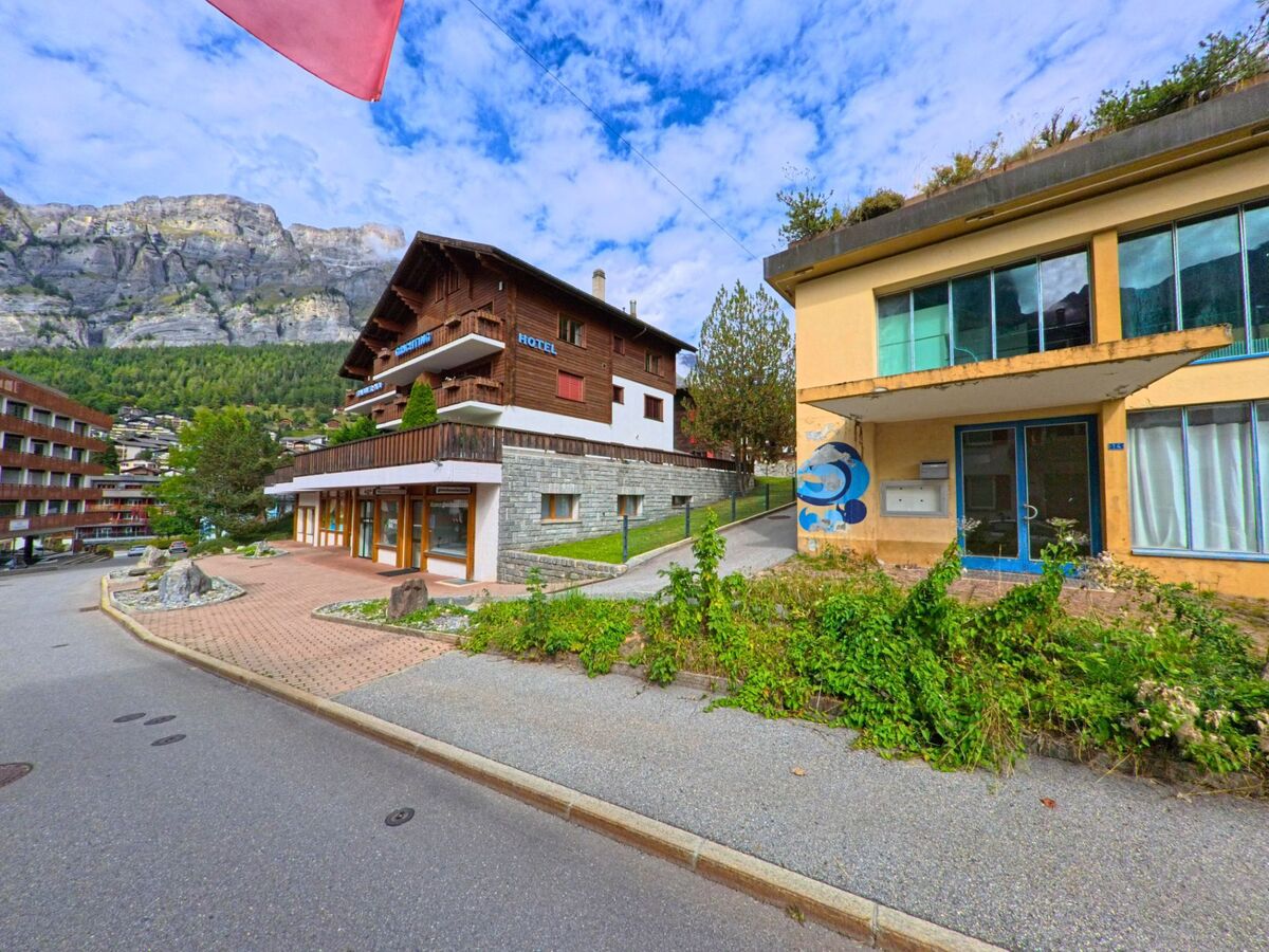 In Leukerbad (3954, Kanton Wallis) steht ein Hotel im Chaletstil mit Holzfassade gegenueber einem aelteren Gebaeude mit grossen Fenstern und sichtbaren Gebrauchsspuren.