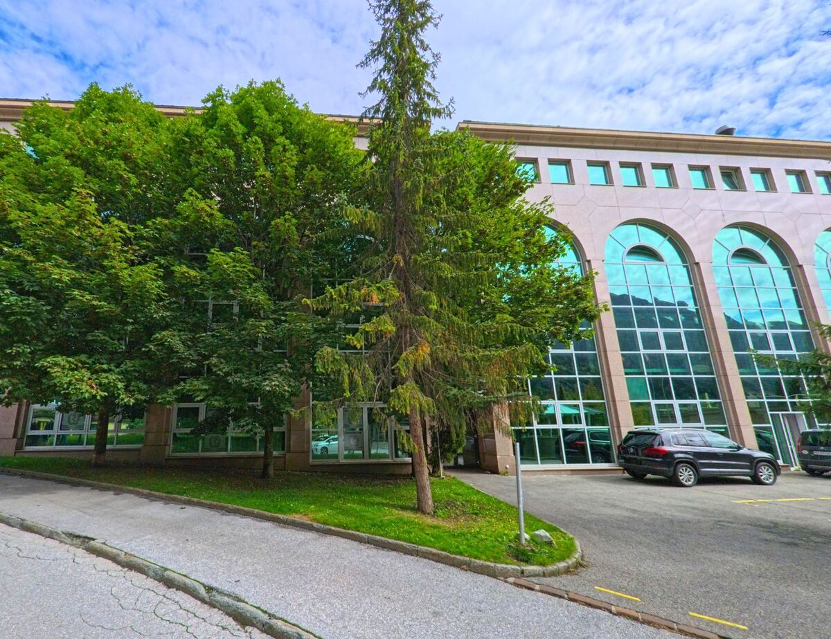 Vor einem modernen Gebaeude mit grossen Glasfassaden in Leukerbad (3954, Kanton Wallis) stehen hohe Baeume, die einen Teil der Ansicht verdecken.