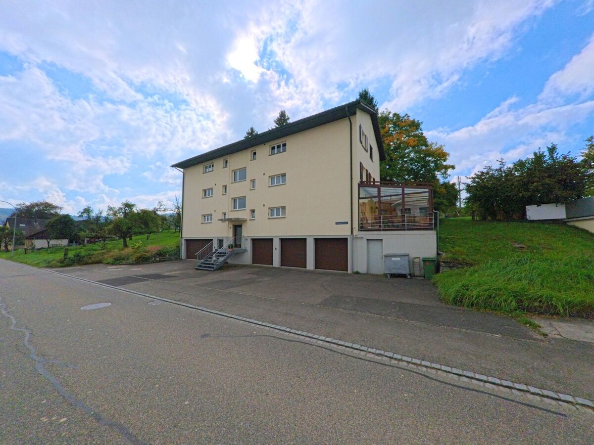 Ein mehrstöckiges Wohnhaus mit Garagen und Wintergarten steht an einer ruhigen Strasse in Leutwil (5725, Kanton Aargau) mit Blick auf grüne Wiesen.