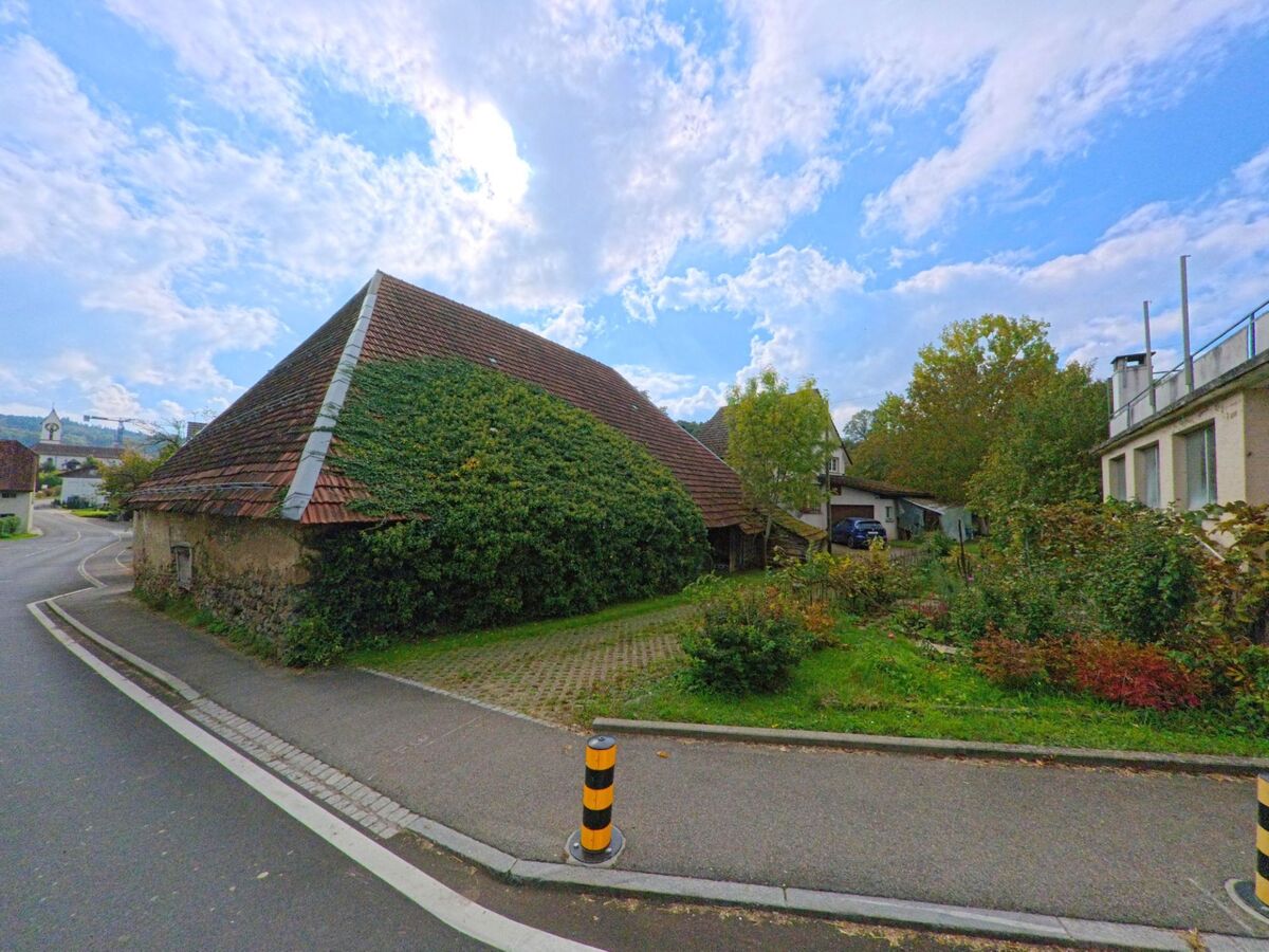 Ein altes, mit Efeu bewachsenes Scheunengebäude steht an einer Kurve in Leutwil (5725, Kanton Aargau) neben einem kleinen Garten und Wohnhäusern.