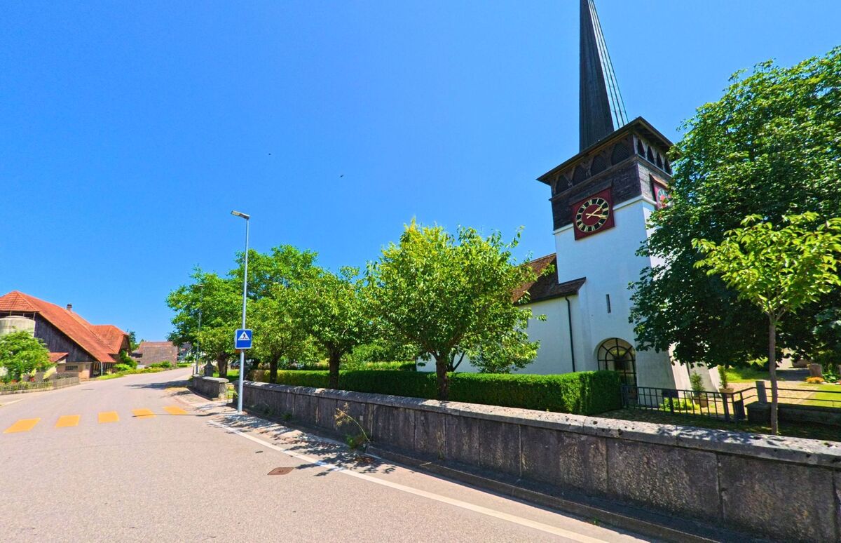 In Leuzigen (3297, Kanton Bern) steht die Kirche mit ihrem markanten Turm und der roten Uhr direkt an einer ruhigen Dorfstrasse mit Fussgängerstreifen.