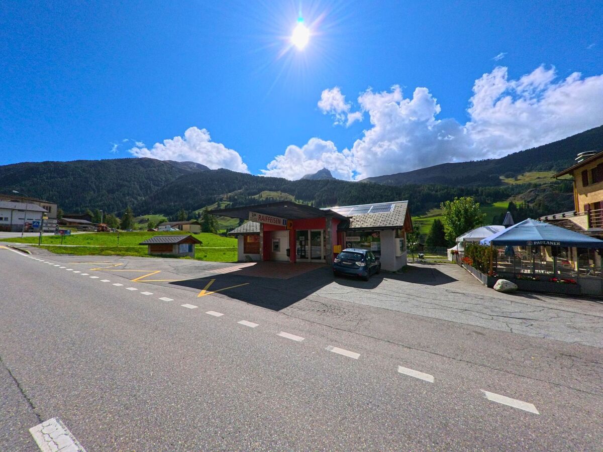 Im Zentrum von Liddes (1945, Kanton Wallis) steht eine kleine Bankfiliale mit Parkplatz, umgeben von Gruenflaechen und Bergen.