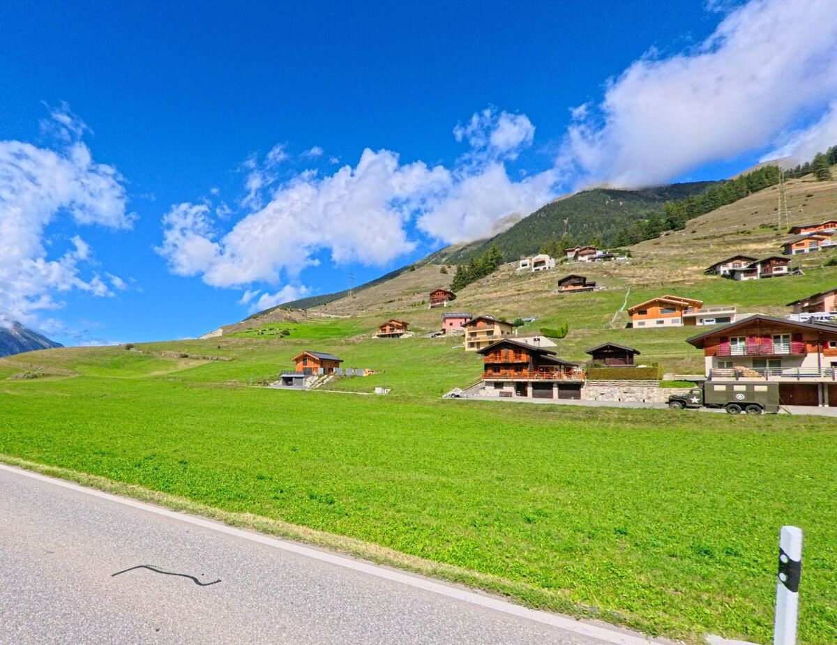 Im Zentrum von Liddes (1945, Kanton Wallis) steht eine kleine Bankfiliale mit Parkplatz, umgeben von Gruenflaechen und Bergen.