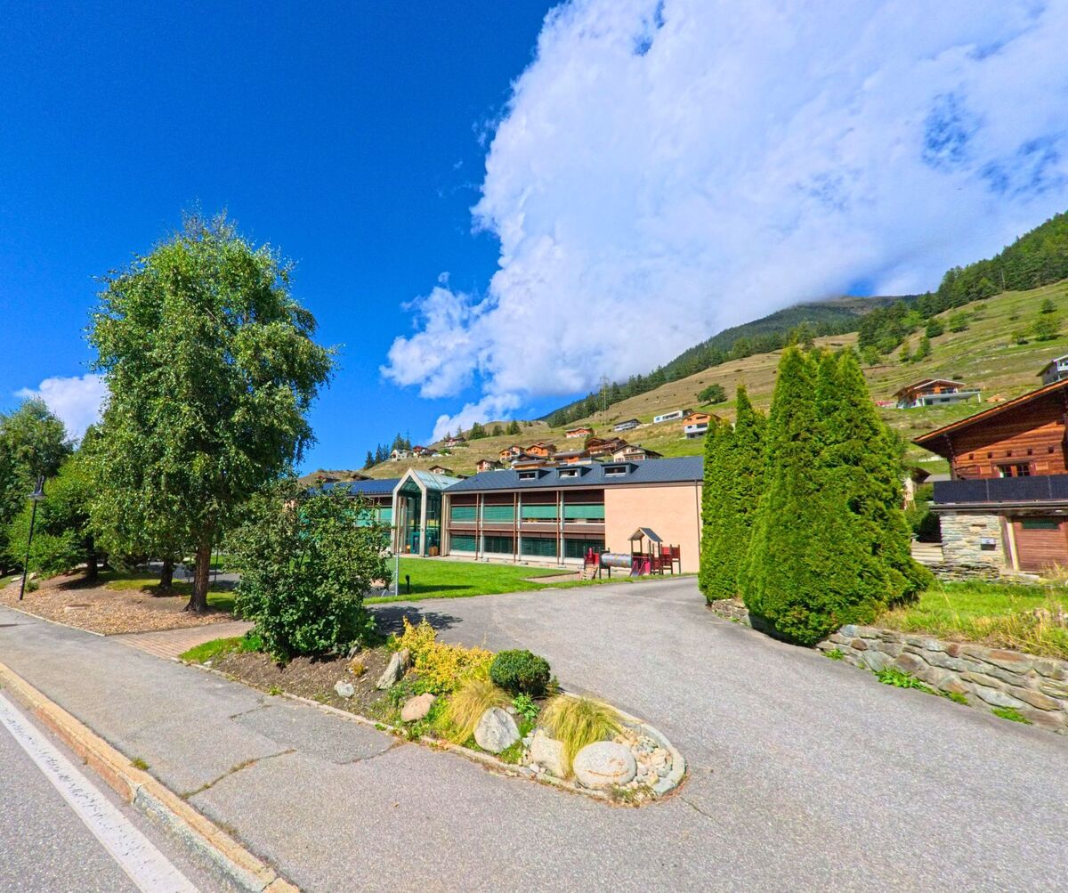 Ein modernes Gebaeude mit grossen Glasfronten steht in Liddes (1945, Kanton Wallis) zwischen traditionellen Chalets am Hang.
