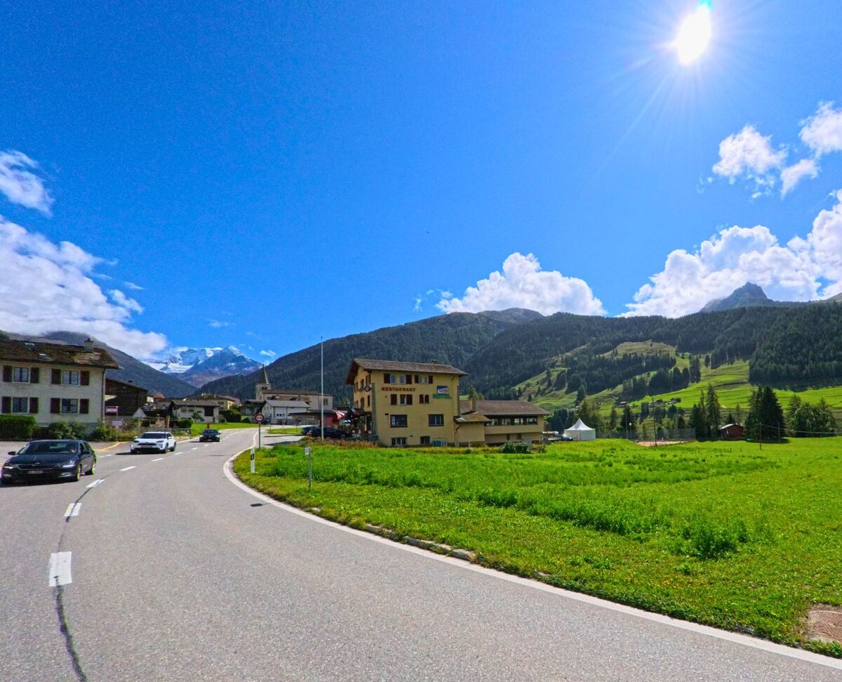 Am Ortseingang von Liddes (1945, Kanton Wallis) stehen mehrere Wohnhaeuser an der Strasse mit Blick auf bewaldete Berge.