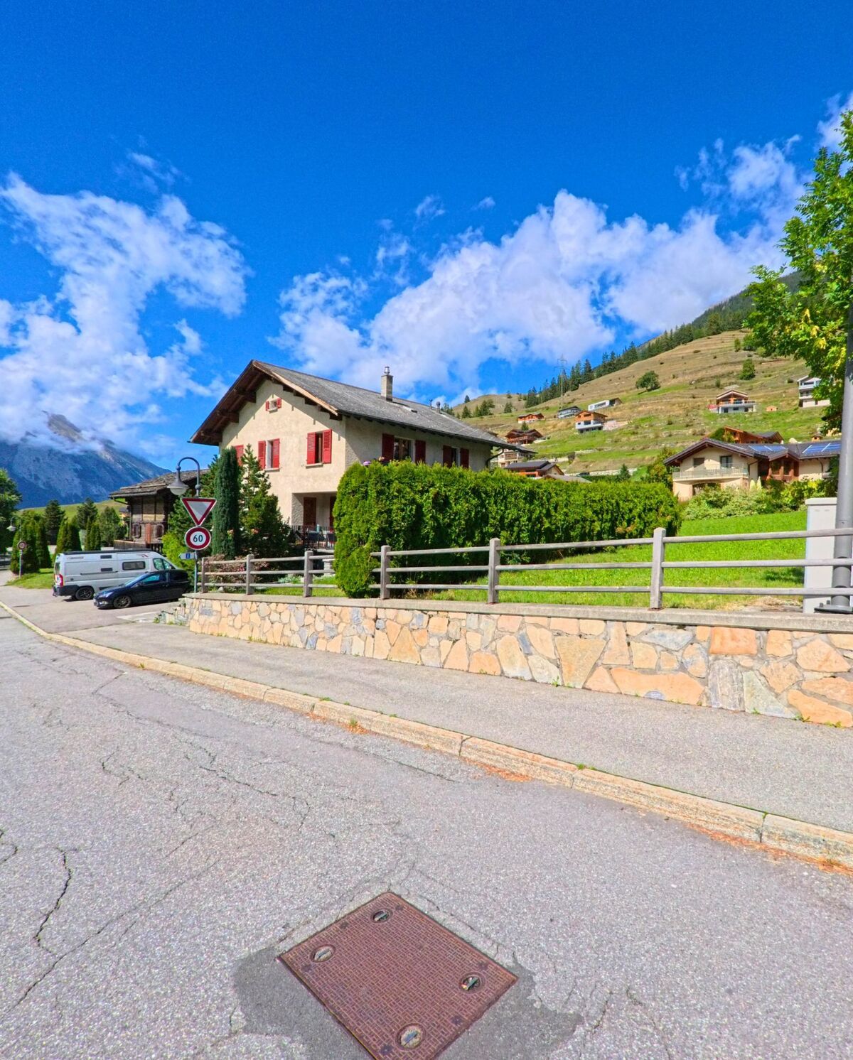 Ein traditionelles Wohnhaus mit roten Fensterlaeden steht in Liddes (1945, Kanton Wallis) am Strassenrand vor sanften Haengen.