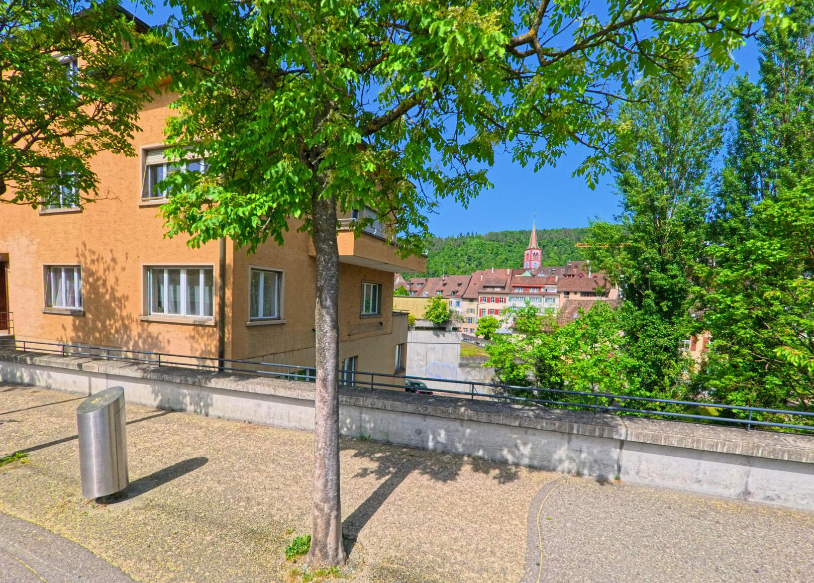 In Liestal (4410, Kanton Baselland) faellt der Blick von einem sonnigen Platz mit Baum und Wohnhaus auf die Dachlandschaft und den Kirchturm der Altstadt.