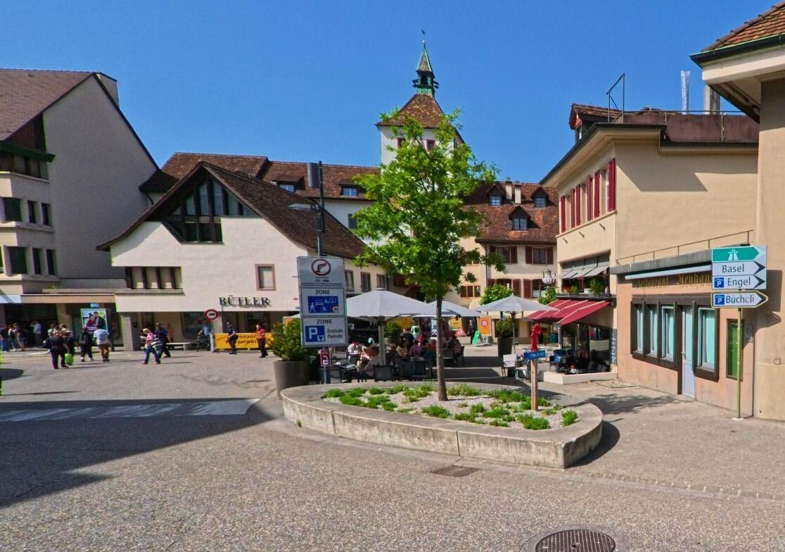 An einer Strassenkreuzung in Liestal (4410, Kanton Basel-Landschaft) steht ein markantes Gebäude mit Turmerker und rotem Ziegeldach vor bewaldetem Hang.