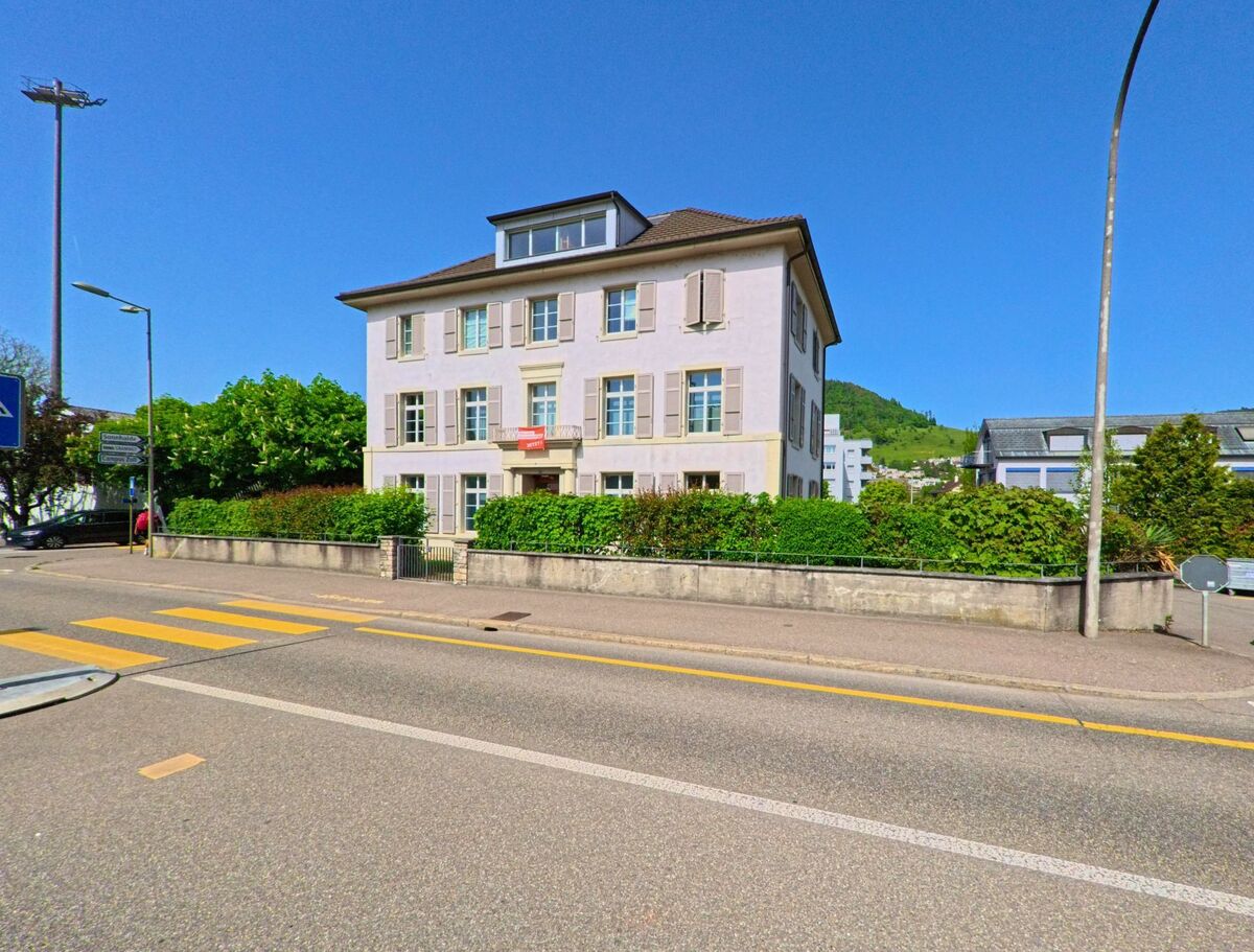 Ein helles Wohnhaus mit Fensterläden und Vorgarten steht an einer Hauptstrasse in Liestal (4410, Kanton Basel-Landschaft) mit Blick auf die umliegenden Hügel.
