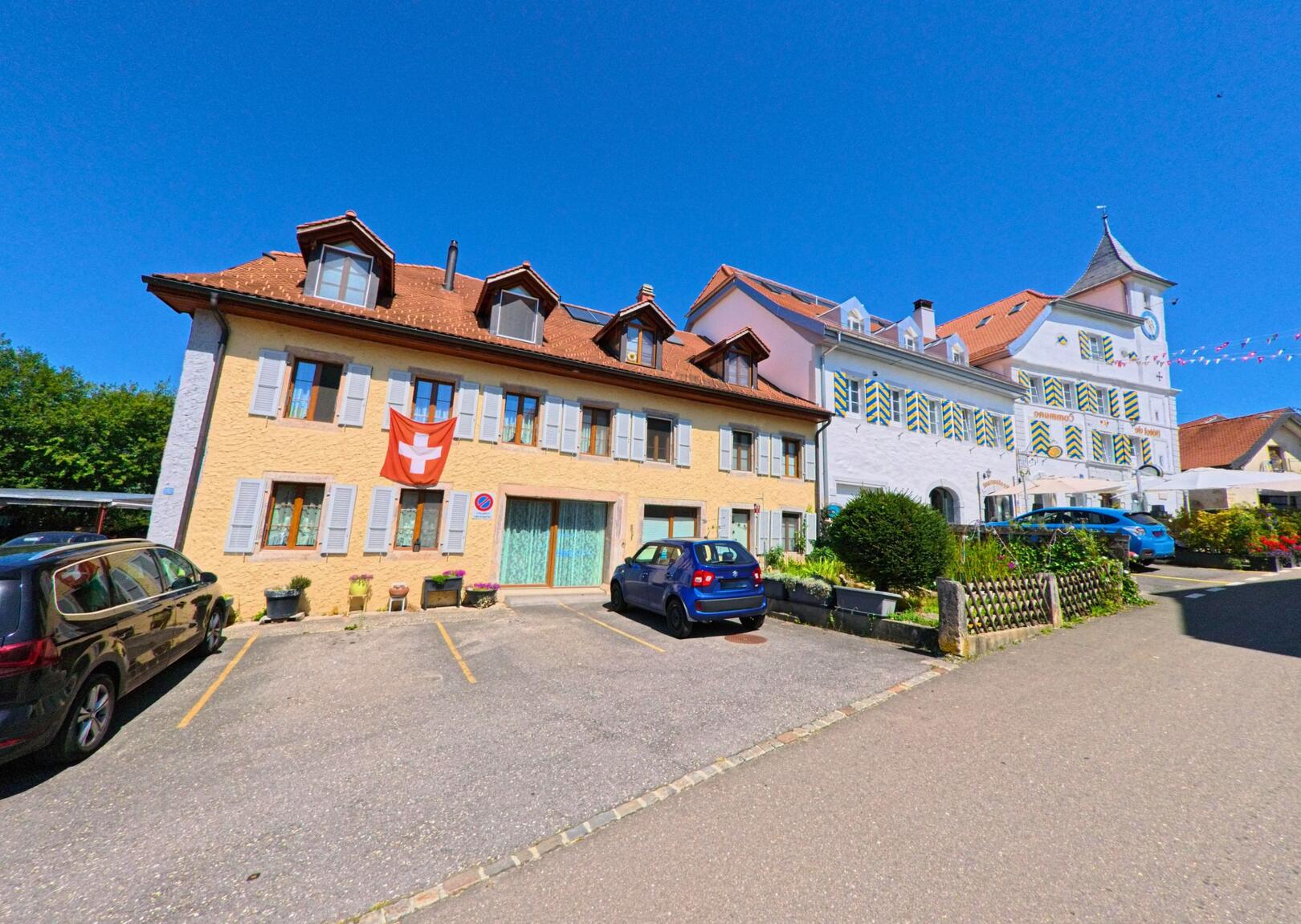 Vor dem markanten weissen Gemeindehaus in Lignieres (2523, Kanton Neuenburg) steht ein gelbes Wohnhaus mit Schweizer Fahne und mehreren geparkten Autos.