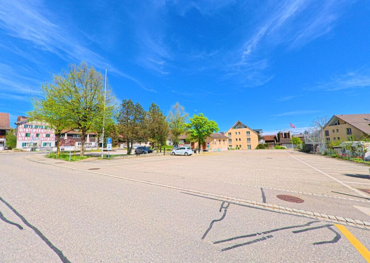 Ein grosser, nahezu leerer Parkplatz in Lindau (8315, Kanton Zürich) wird von Bäumen und verschiedenen Wohn- sowie Fachwerkhäusern umrahmt, im Hintergrund ist eine Fahne zu sehen.