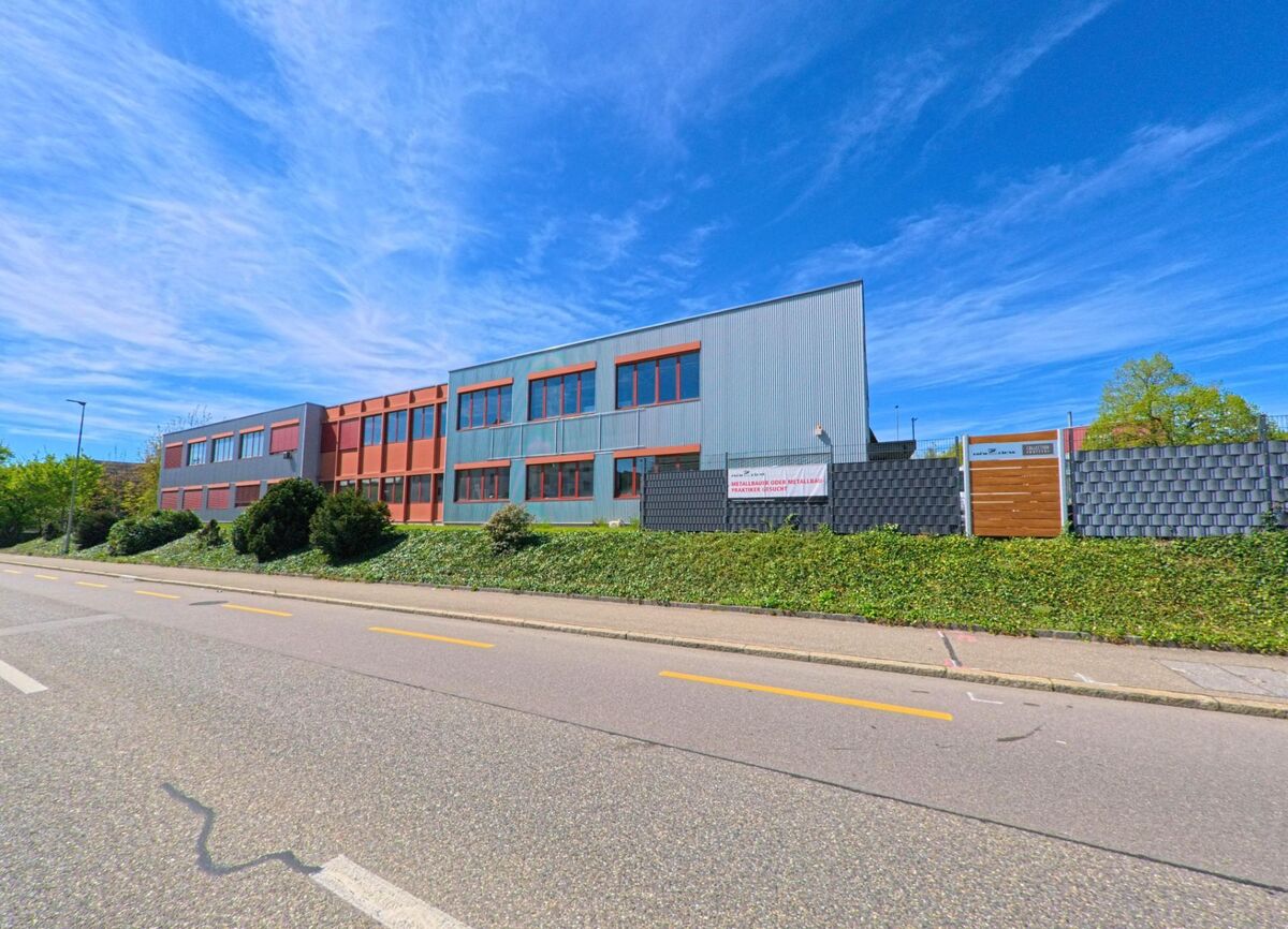 An einer Hauptstrasse in Lindau (8315, Kanton Zürich) steht ein modernes Industriegebäude mit grossen Fenstern und grauen Fassadenplatten, umgeben von einer gepflegten Hecke.