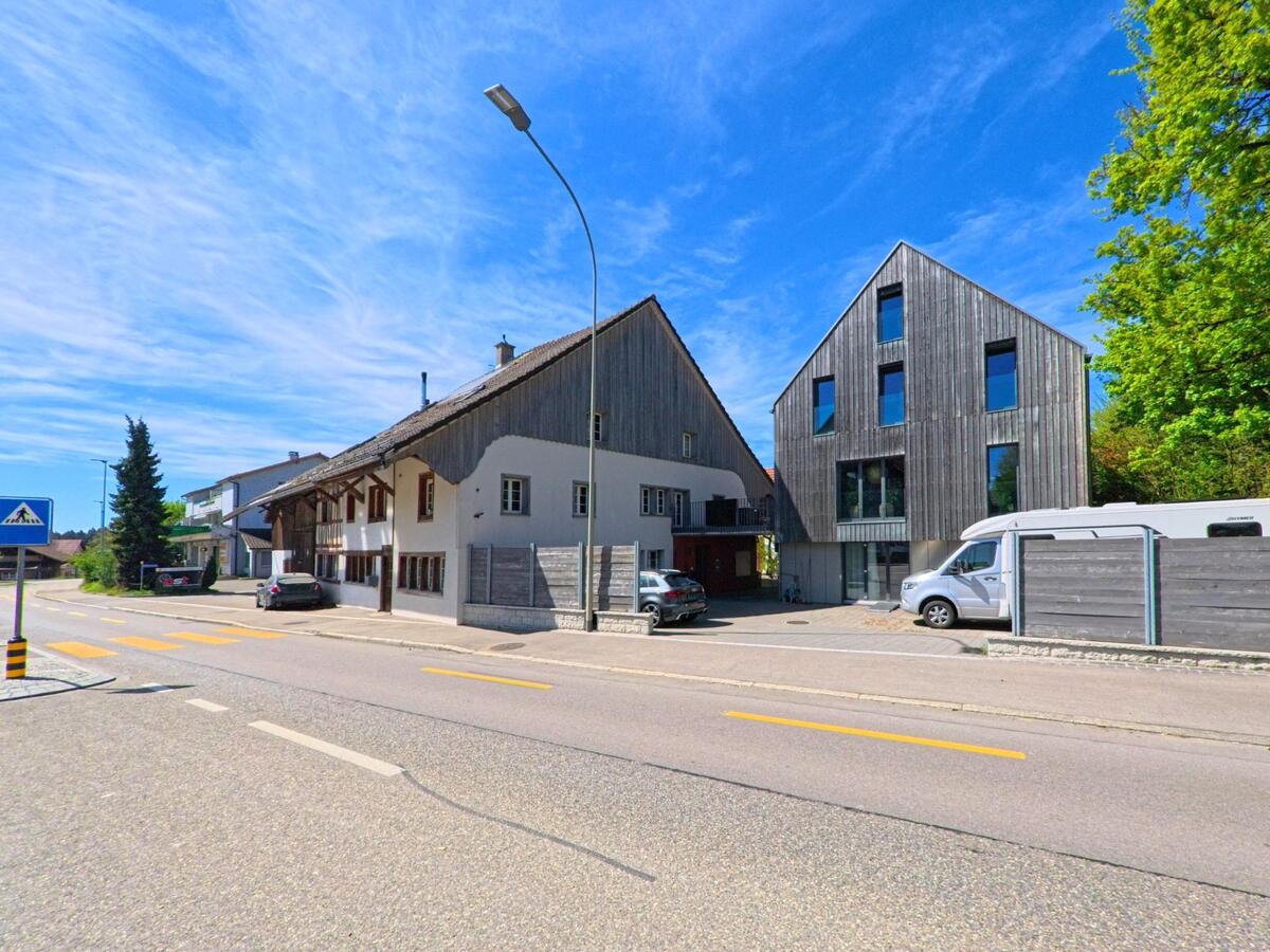 An einer Strasse in Lindau (8315, Kanton Zürich) stehen ein traditionelles Wohnhaus und ein modernes Holzgebäude nebeneinander, im Vordergrund ist ein Zebrastreifen zu sehen.